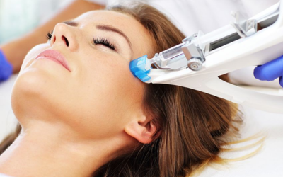 Woman receiving a professional facial treatment; a clinician with blue gloves uses a handheld device on her cheek.