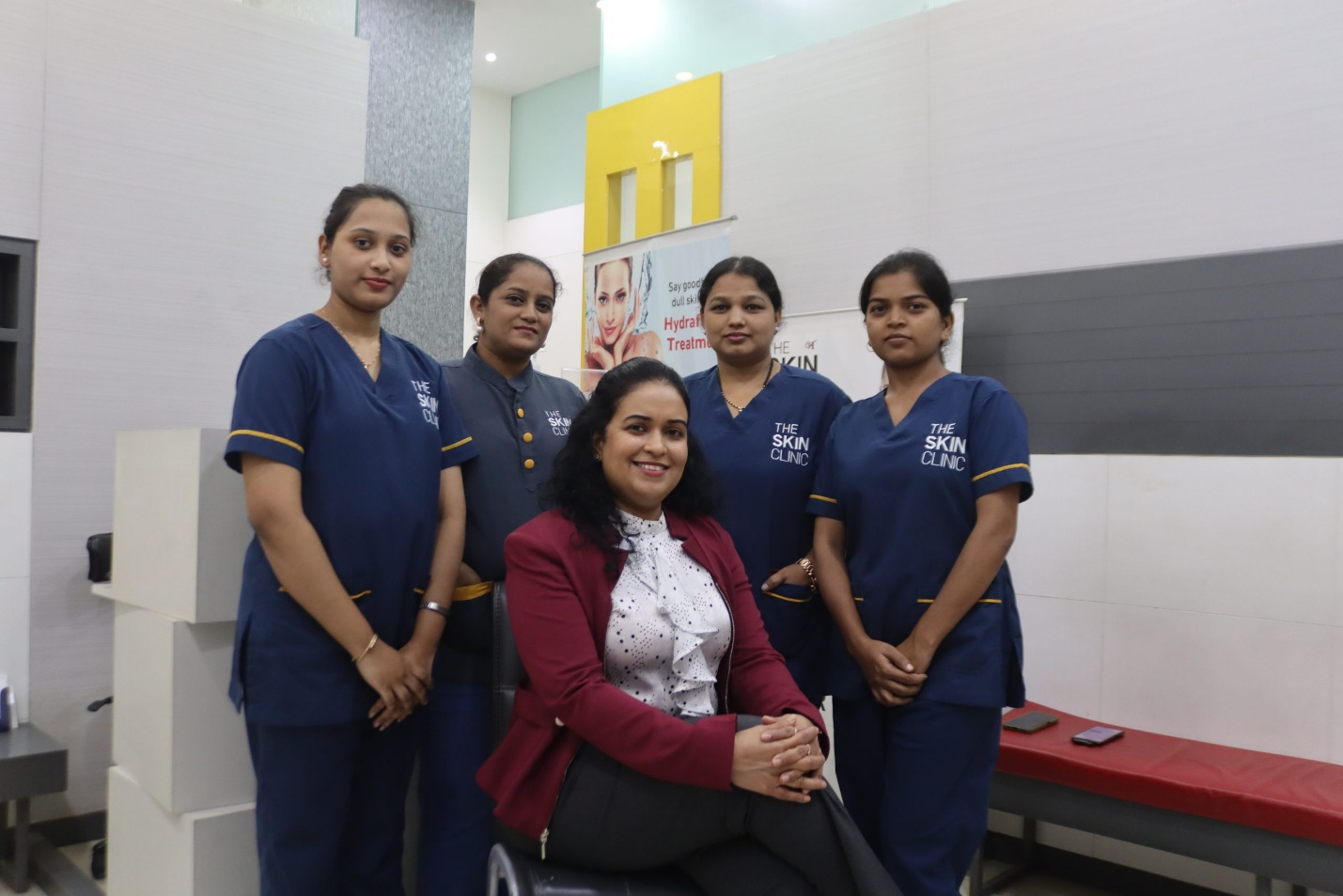Picture3(1) Group portrait of five women inside a skin clinic: four staff in navy scrubs stand behind a seated client in a burgundy blazer, all smiling.