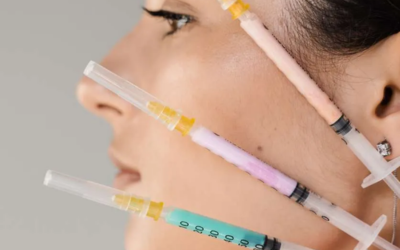 Side profile of a woman receiving facial injections with three syringes near her cheek, containing yellow, pink, and blue liquid.