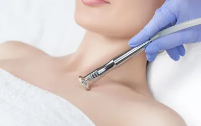 Clinician with a blue glove uses a handheld cosmetic device on a woman's bare neck during a treatment.